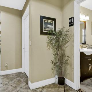 A hallway with tile flooring, featuring a potted plant and artwork on the wall, leading to a bathroom with a mirror and vanity.