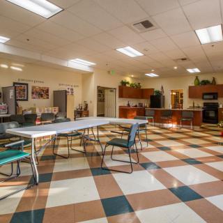 A spacious community room with a kitchen, featuring tables and chairs on a checkered floor.