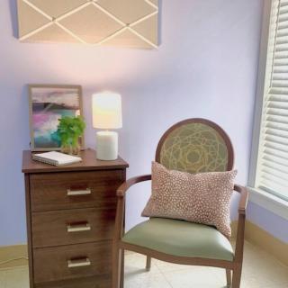 A cozy corner with a wooden chair and cushion, next to a small desk featuring a lamp and decor.