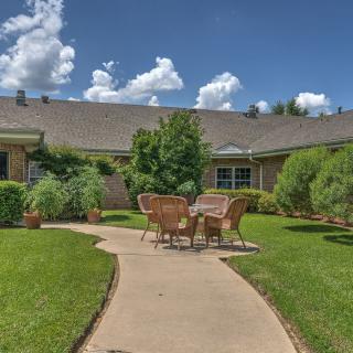 A scenic garden with a pathway leading to patio furniture surrounded by lush greenery and under a clear blue sky.