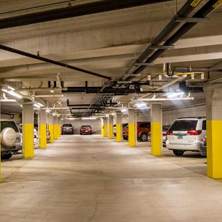 An underground parking garage with several cars parked alongside concrete pillars under fluorescent lights.