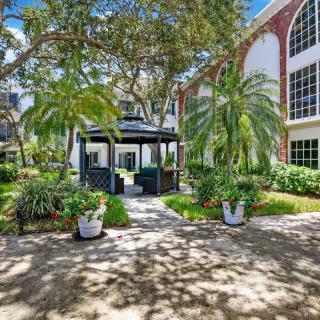 A peaceful garden with a gazebo surrounded by palm trees and greenery in front of a large building.