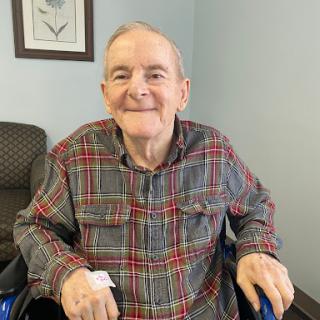 A cheerful elderly man sitting in a wheelchair, dressed in a plaid shirt, enjoying his time indoors.