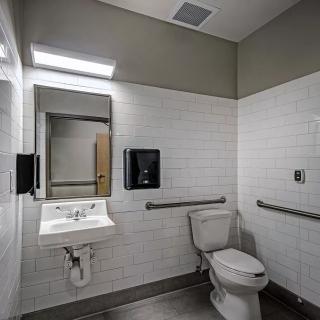 A clean and accessible bathroom with white tiled walls, featuring a toilet, sink, mirror, and grab bars.