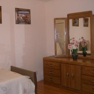 A cozy bedroom featuring a wooden dresser with mirrors and decorative items, alongside a bed and wall art.