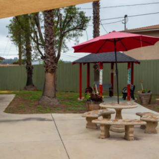 A patio area with a stone table, benches, and a red umbrella, surrounded by trees and a green fence.
