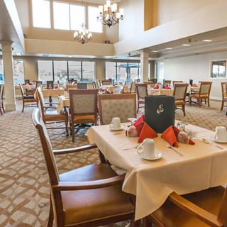 A spacious dining area with elegantly set tables and comfortable chairs, featuring large windows and chandeliers.