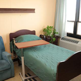A well-lit hospital room featuring a single bed with green bedding, a comfortable blue chair, and a small table for patient needs.