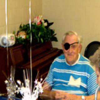 An elderly man wearing an eye patch sits at a table with balloons and a piano in the background.