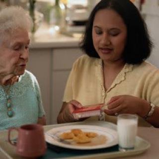 A caregiver assists an elderly woman with her meal in the kitchen.