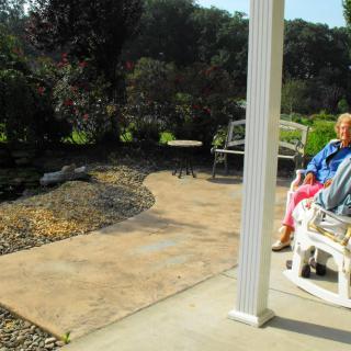 An elderly person sits on a patio bench on a sunny day, overlooking a garden path.