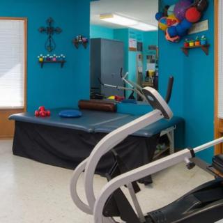 A small indoor gym with exercise equipment including a treadmill, therapy table, and various fitness accessories.