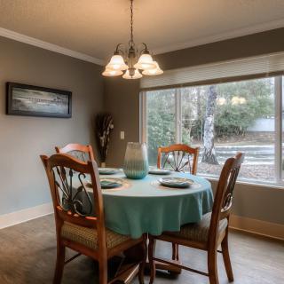 A cozy dining room with a round table set for four, illuminated by a hanging chandelier and bathed in natural light from a large window.
