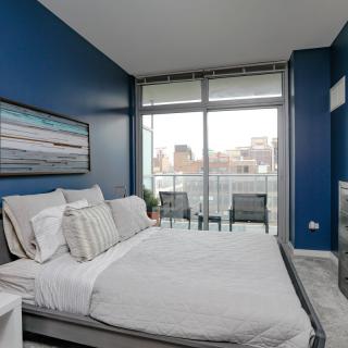 A modern bedroom with blue walls, featuring a neatly made bed and large windows leading to a balcony.