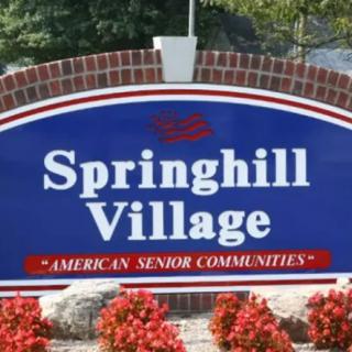 A sign for Springhill Village, an American Senior Communities residence, displayed with a brick base and flowers.