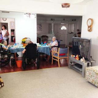 A cozy living room with elderly individuals gathered around a table, some in wheelchairs, with a television and home decor visible.