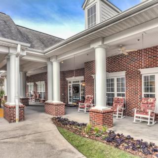 An elegant brick building with a porch featuring white columns and rocking chairs.