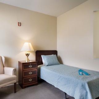 A minimalist bedroom featuring a single bed, a nightstand with a lamp, and an upholstered chair.