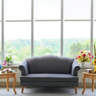 A cozy living room setup with a sofa, flanked by coffee tables and vibrant flower arrangements in front of large windows.