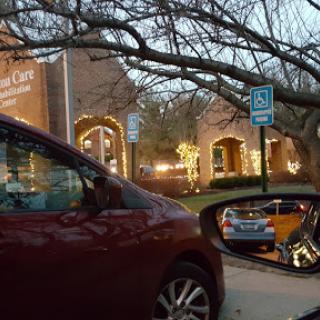A red car parked in a lot, reflecting on the side mirror, with a brightly lit building and tree branches visible in the evening.