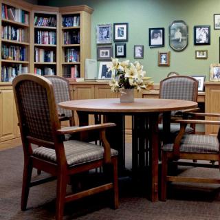 A cozy library meeting area with a round table surrounded by chairs, bookshelves filled with books, and a wall adorned with framed photos.