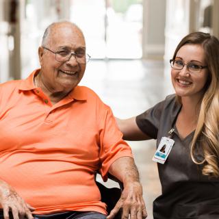 An elderly person in a wheelchair smiling with a caregiver inside a healthcare facility.