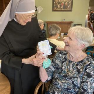 An elderly woman smiles as a nun hands her a card, showcasing joyful interaction in a nursing home setting.