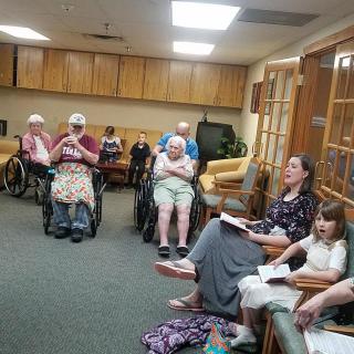 A group of elderly individuals and a young girl participate in a community gathering at a nursing home.