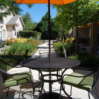 A serene outdoor patio with a table, chairs, and an orange umbrella amidst lush greenery.