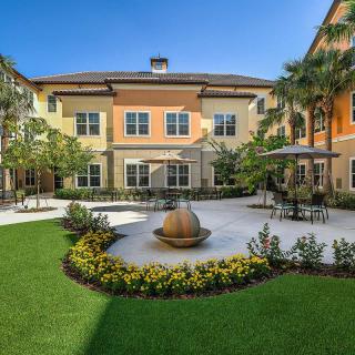 A beautifully landscaped courtyard featuring a central fountain, surrounded by a building with outdoor seating and palm trees.