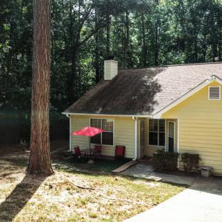 A charming yellow house nestled among tall trees with a red umbrella on the patio.