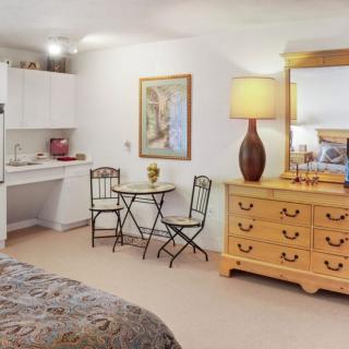 A cozy bedroom interior featuring a dresser with a lamp, a small table with chairs, and wall artwork.