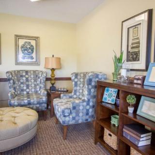A cozy living room with patterned chairs, an ottoman, and a bookshelf filled with books and decor.