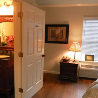 A cozy bedroom with a nightstand and a lamp next to the window, leading into an adjacent room.