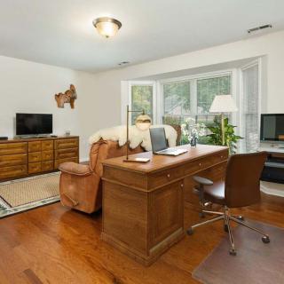 A cozy home office space with a wooden desk and chair, adjacent to a living area featuring a sofa, television, and large windows overlooking greenery.