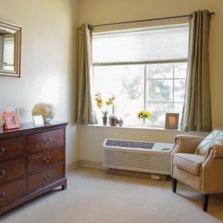 A cozy bedroom featuring a dresser, an armchair by the window, and decorative items arranged on the furniture.