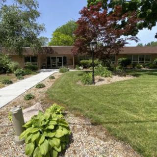 A landscaped area with a building in the background, featuring a sidewalk, trees, and well-maintained greenery.