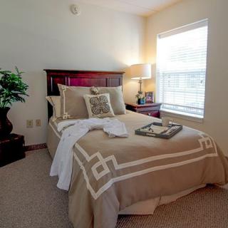 A cozy bedroom featuring a neatly made bed with decorative pillows, a bedside lamp, and a large window allowing natural light.