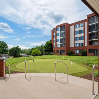 A view of a putting green surrounded by trees and a multi-story building under a partly cloudy sky.
