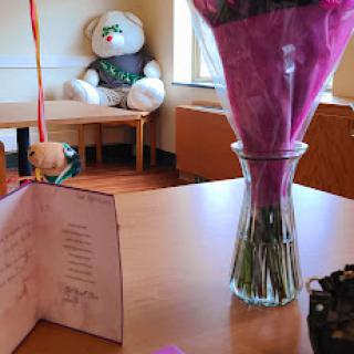 A beautifully arranged bouquet in a vase next to a heartfelt greeting card, with a large teddy bear in the background.