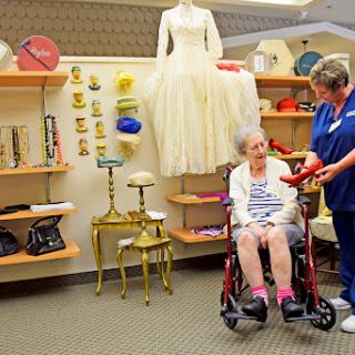 An elderly woman in a wheelchair is being assisted by a caregiver while browsing a display of vintage clothing and accessories.