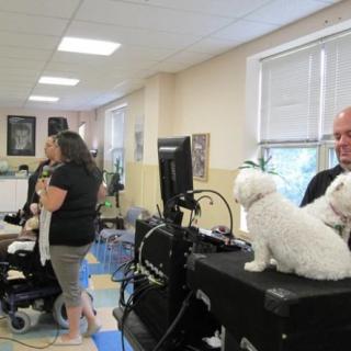 A community event taking place indoors, featuring people interacting with two white dogs and using a microphone for announcements.