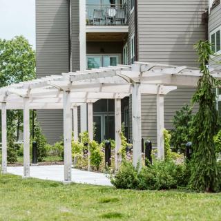 A white pergola in front of a modern apartment building, surrounded by lush greenery.
