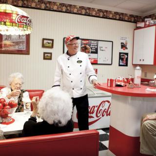 A retro-style diner with a chef speaking to patrons, surrounded by Coca-Cola themed decor.
