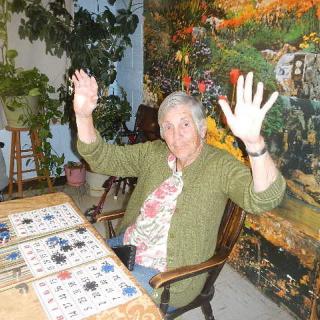 An elderly woman raises her hands in celebration while playing bingo indoors.
