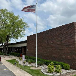 A view of a brick building with an American flag flying in front, surrounded by landscaped greenery on a cloudy day.