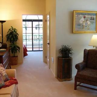 A cozy living room with comfortable seating, artwork on the wall, and indoor plants, leading to a bedroom.