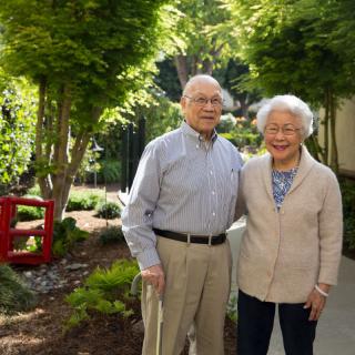 An elderly couple standing together in a sunny garden.
