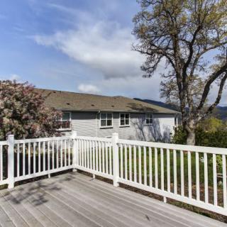 A spacious wooden deck with white railings overlooks a yard with trees and a house in the background under a partly cloudy sky.