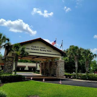 The entrance of a facility, featuring a welcoming sign and flags against a clear blue sky.